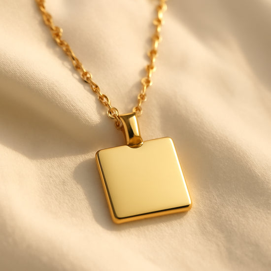 A close‑up of a gold square pendant necklace resting on a soft white fabric, with warm natural light highlighting its polished edges. Alt: Classic gold square pendant necklace highlighting elegant geometry and shine.