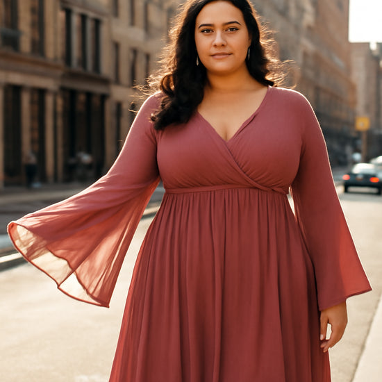 A model in a flowing empire‑waist dress with bell sleeves standing on a city street, sunlight catching the gentle flare of the sleeves. Alt: Plus size evening dress with bell sleeves in a sunny city setting.